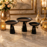 Home Sajawat set of three matte black pedestal serving stands with gold branch and butterfly accents on a marble table