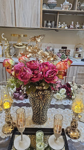 Home Sajawat artificial roses and peonies in a textured gold vase centerpiece on a dining table with candle holders.