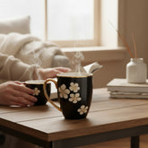 Home Sajawat black ceramic mug with white floral pattern and gold handle, steaming on a wooden table.