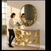 Woman styling a Home Sajawat gold metal floral console table and matching round leaf-adorned wall mirror in a hallway.