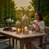 Home Sajawat ornate metal candle lanterns with filigree patterns on a wooden patio table with a woman drinking tea.