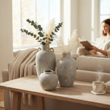 Home Sajawat set of three grey-blue textured ceramic amphora vases with gold handles and rims, holding dried pampas grass and eucalyptus.