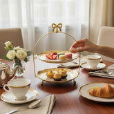 Home Sajawat two-tiered serving stand with gold-rimmed textured plates and a decorative bow handle on a dining table.