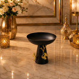 Home Sajawat matte black pedestal bowl with gold ginkgo leaf accents on a marble table with warm ambient lighting.