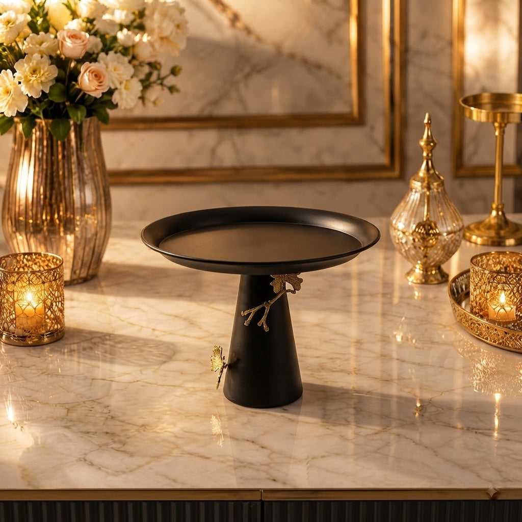 Home Sajawat matte black pedestal serving stand with gold branch and butterfly accents on a marble table.