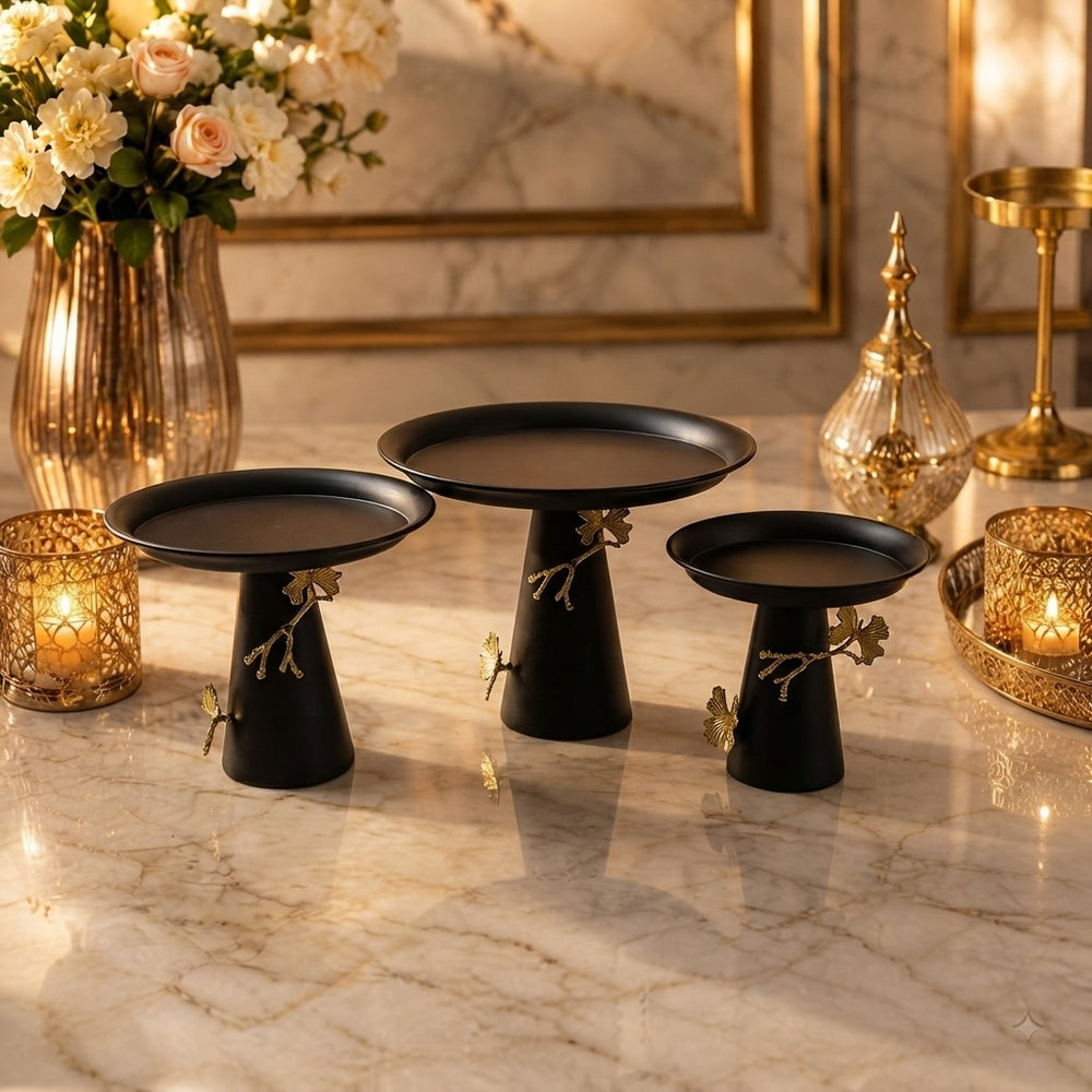 Home Sajawat set of three matte black pedestal serving stands with gold branch and butterfly accents on a marble table