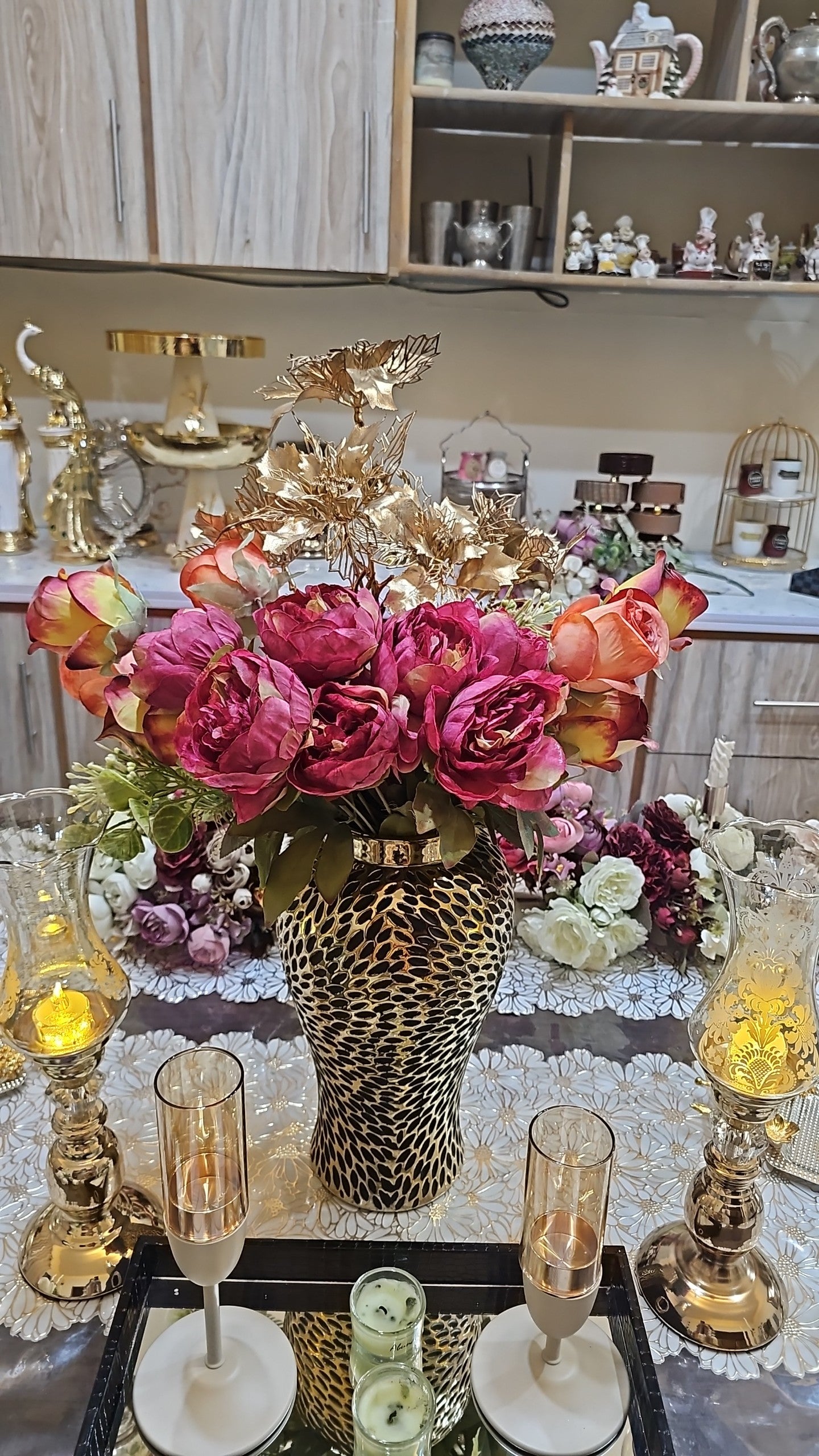 Home Sajawat artificial roses and peonies in a textured gold vase centerpiece on a dining table with candle holders.