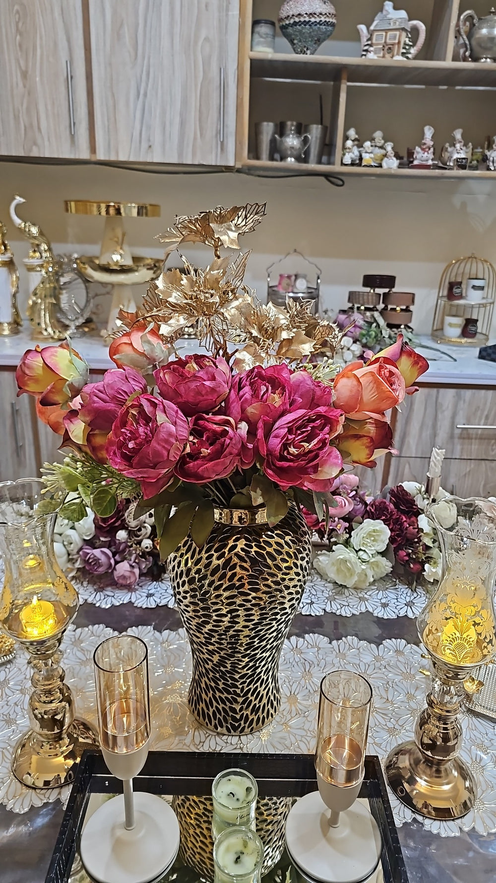 Home Sajawat artificial roses and peonies in a textured gold vase centerpiece on a dining table with candle holders.