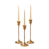 Home Sajawat set of three gold metal candlesticks with cream taper candles on a dining table.