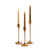 Home Sajawat set of three gold metal taper candle holders with varying heights, holding cream, blush, and olive green candles.