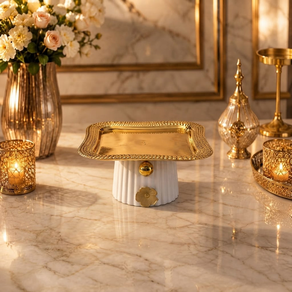 Home Sajawat square pedestal serving tray with a polished gold metal top and a white fluted ceramic base.