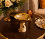 Home Sajawat decorative pedestal bowl with a hammered gold-tone finish and a cream ceramic base with ginkgo accents.