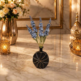 Home Sajawat matte black circular vase with intricate carved details holding blue faux floral stems on marble table