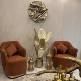 Home Sajawat decorative metal leaf floor sculpture in brushed gold and white, placed between two rust-orange chairs.