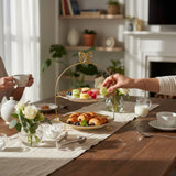Home Sajawat two-tiered serving stand with gold-rimmed plates and decorative bow handle, styled on a dining table.